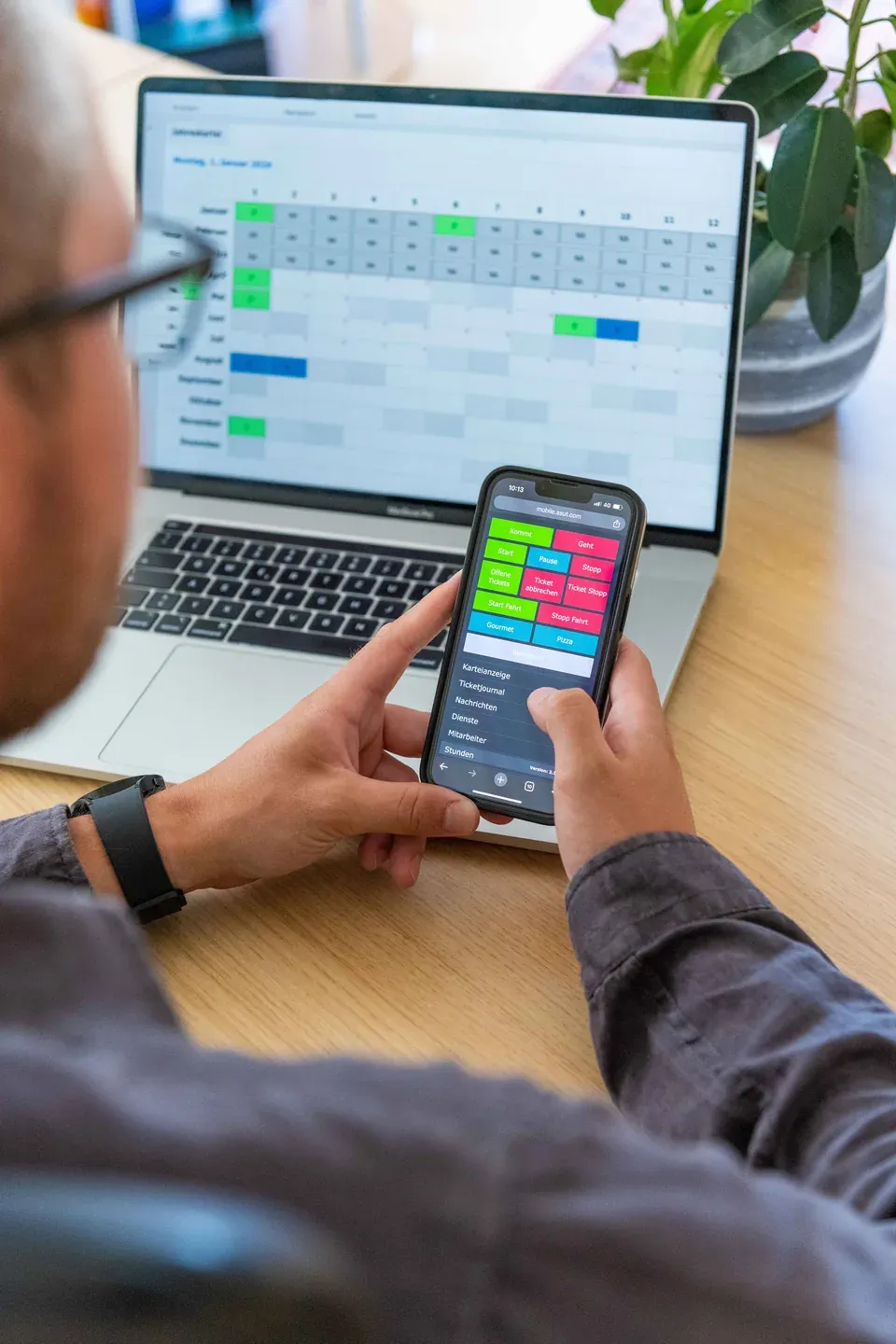 Timework Stempel-App am Smartphone mit Kommt- und Geht-Buttons, im Hintergrund die Jahreskartei am Laptop