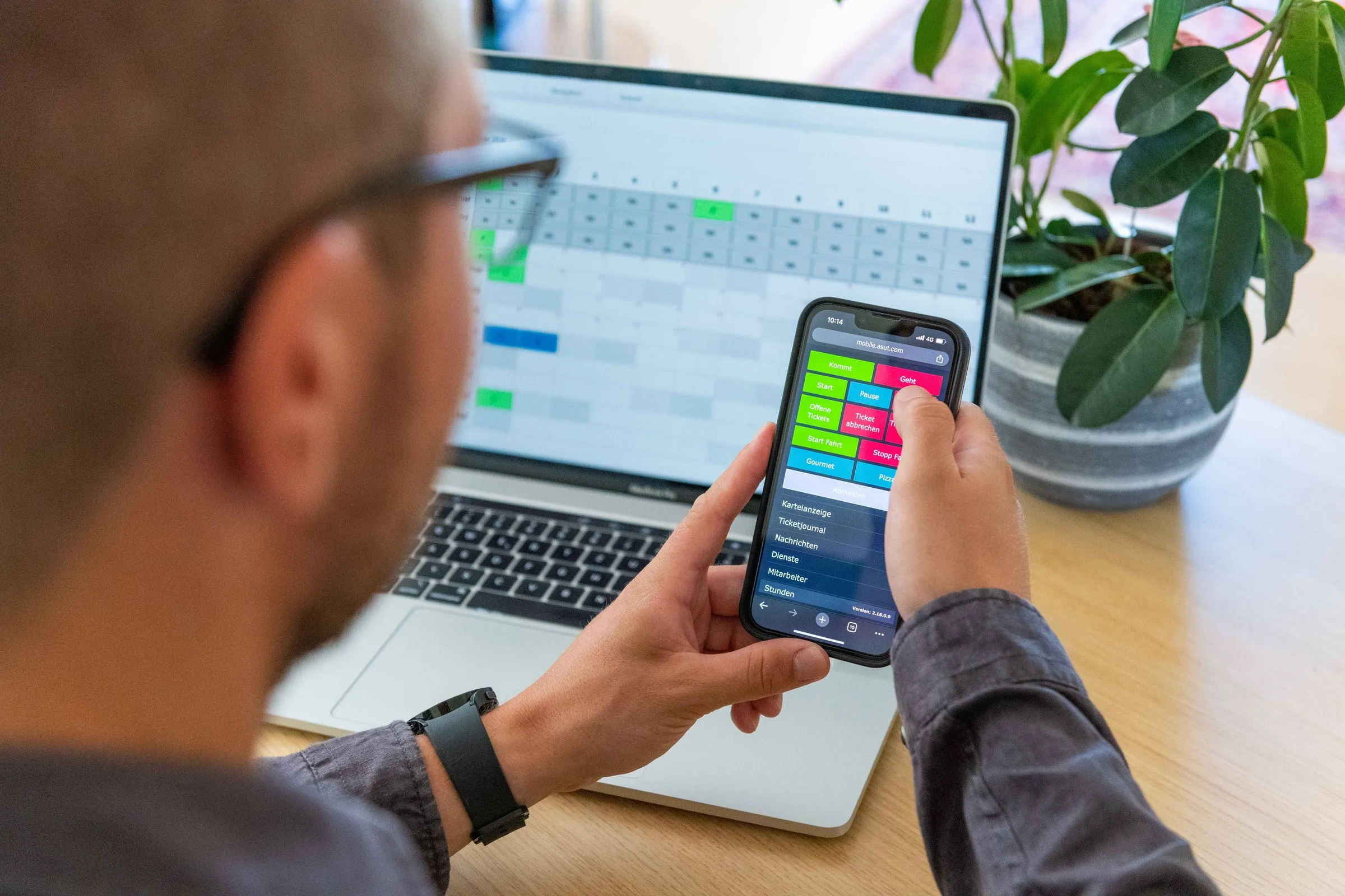 Mitarbeiter bucht Projekt- und Auftragszeit in der Timework-App am Smartphone, im Hintergrund die Auswertung am Laptop