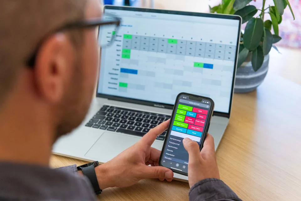 Mobile Zeiterfassung mit der Timework-App am Smartphone, im Hintergrund die Jahreskartei am Laptop
